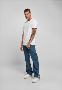 Witte katoenen t-shirt, blauwe denim jeans en witte sneakers. Model toont een tatoeage op de linkerarm. Eenvoudige snit en relaxte pasvorm.