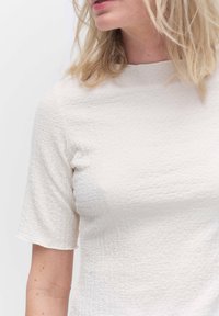 Teksturowany, off-white top z krótkimi rękawami i wysokim dekoltem. Materiał ma subtelny wzór marszczony, charakteryzujący się dopasowanym krojem.