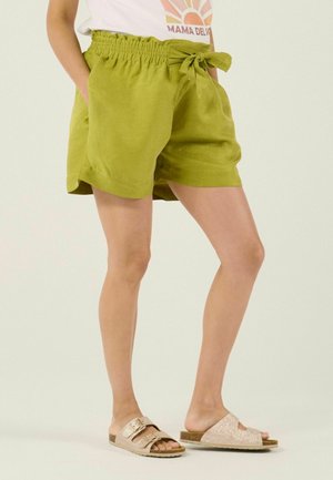 Femme portant un short taille haute vert citron avec un nœud, un t-shirt blanc à motif graphique et des sandales beige à enfiler avec des boucles.