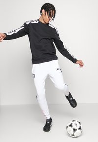Schwarzer Sweatshirt mit weißen Streifen, weiße Hose mit schwarzen Akzenten und schwarze Fußballschuhe. Die Person spielt mit einem schwarz-weißen Fußball.