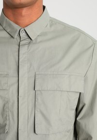 Zbliżenie osoby w jasnoszarej koszuli z guzikami, z dwiema kieszeniami na klatce piersiowej i z wyłogami w kształcie ostrym.