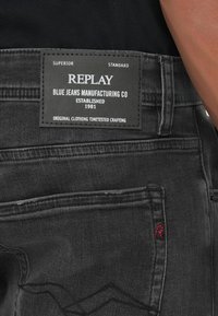 Crne traperice od denim materijala sa pravokutnom etiketom na kojoj piše "Replay Blue Jeans Manufacturing Co" i crvenim šavom u blizini džepa.