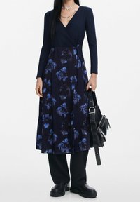 Vestido envolvente de color navy con mangas largas, patrón floral en tonos de azul, falda fluida, combinado con un bolso negro y zapatos gruesos.