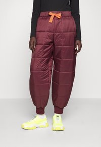 Bordeauxfarbene, gesteppten Hosen mit oranges Zugband, ausgestattet mit elastischen Bündchen und einer lockeren Silhouette. Kombiniert mit leuchtend gelben Turnschuhen.