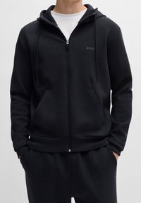 Svart zip-up hoodie i mjukt tyg med en framficka, dragskor och en subtil logotyp på bröstet; ihop med mörka byxor.