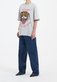 Šedé oversized tričko s barevným grafickým motivem tygra, který obsahuje černé, žluté a červené detaily, v kombinaci s volnými modrými džíny a špičatými černými botami.