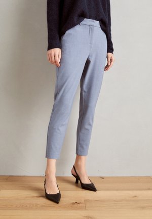 Osoba ubrana w dopasowane, jasnoniebieskie spodnie o długości do kostek, czarne czółenka na obcasie z odkrytą piętą i czarną bluzkę z długim rękawem, stojąca na drewnianej podłodze.