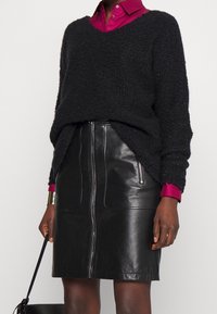 Sort tekstureret sweater med v-udskæring og lange ærmer lagdelt over en fuchsia skjorte med krave. En sort lædernederdel med en lynlås foran.