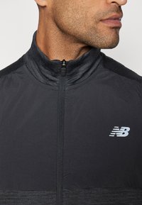 Tuvojoties vīrietim, kurš valkā melnu sporta jakas ar rāvējslēdzēju un augstu apkakli, uz krūtīm ir balts New Balance logo.