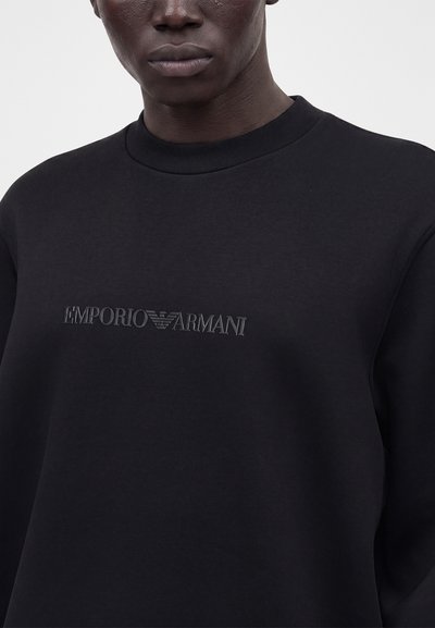 Personne à la peau foncée portant un sweat-shirt noir avec le logo "EMPORIO ARMANI" en lettres noires embossées sur la poitrine.
