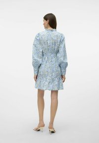 Blomstrete kjole i lys blå med samlet midje og flosset kant. Lange, puffete ermer og subtil glans. Hæler synlige.