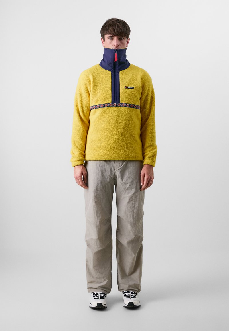 Geel fleece pullover met ritssluiting en marineblauwe kraag, voorzien van een bloemenpatroon band, gecombineerd met lichtgrijze broek en zwart-witte schoenen.