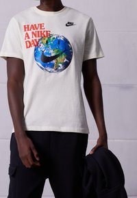 Vaalean kerma valkoinen puuvillapaita, jossa on värikäs maapallon grafiikka hymyilevällä kasvoilla ja Nike-logo; lyhyet hihat, rento istuvuus.
