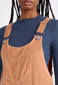 Lichtbraune Corduroy-Latzhose mit einer vorderen Tasche, ausgestattet mit Metallclipverschluss und verstellbaren Trägern, getragen über einem gerippten navy-blauen Shirt.