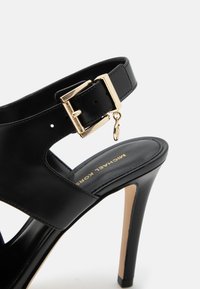 Sandale à talon haut en cuir noir avec boucle dorée et petit pendentif suspendu, présentant un design ajouré et une semelle intérieure de marque.