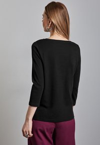 Schwarzes Langarmshirt mit einem U-Boot-Ausschnitt, aus weichem Material gefertigt. Verfügt über Dreiviertelärmel und einen lockeren Schnitt. Getragen mit bordeauxfarbenen Hosen.
