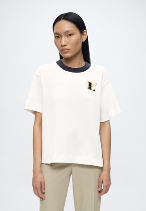 Joven mujer con cabello largo y negro lleva una camiseta blanca de manga corta con cuello negro y una "L" bordada en el pecho, combinada con pantalones beige.