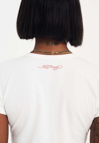 Witte katoenen t-shirt met korte mouwen. Achterkant met roze geborduurde tekst "Ed Hardy." Ketting halsketting zichtbaar aan de achterkant van de nek.