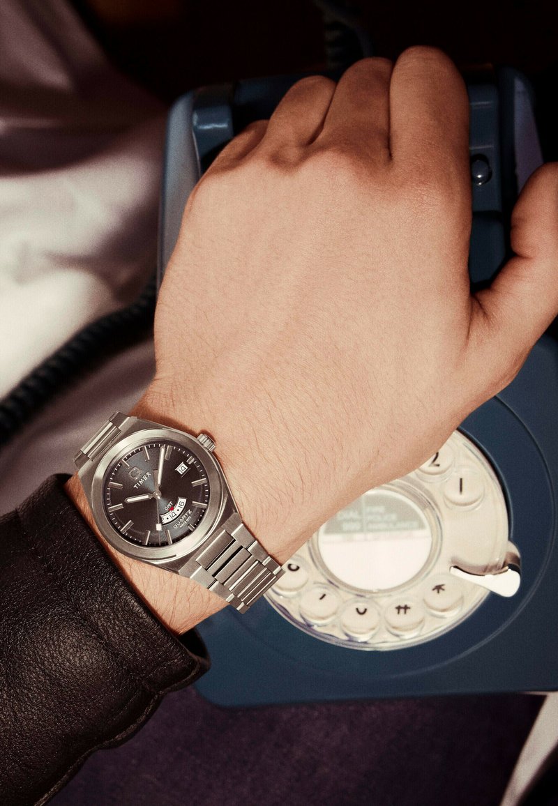 Hand mit einer Edelstahl-Timex-Uhr, die eine Nummer auf einem blauen Wählscheiben-Telefon wählt, mit sichtbarem Ärmel des Mantels.