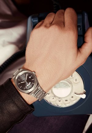 Main droite portant une montre en argent tenant le récepteur d'un téléphone à cadran rotatif bleu sur une surface sombre.