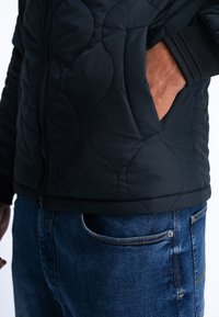 Schwarze gesteppte Jacke mit einem strukturierten Muster, mit einem Reißverschluss vorne und gerippten Bündchen. Getragen über blauen Jeans mit sichtbaren Taschennähten.