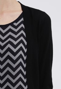 Cardigan nero aperto davanti indossato sopra una camicia blu navy con strisce bianche a chevron, che mette in risalto una texture liscia e un design classico.