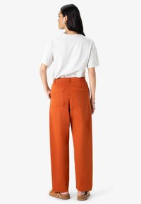 Pantalons orange à jambes larges avec poches arrière, accompagnés d'un t-shirt gris clair à manches courtes. Tissu lisse et coupe décontractée.