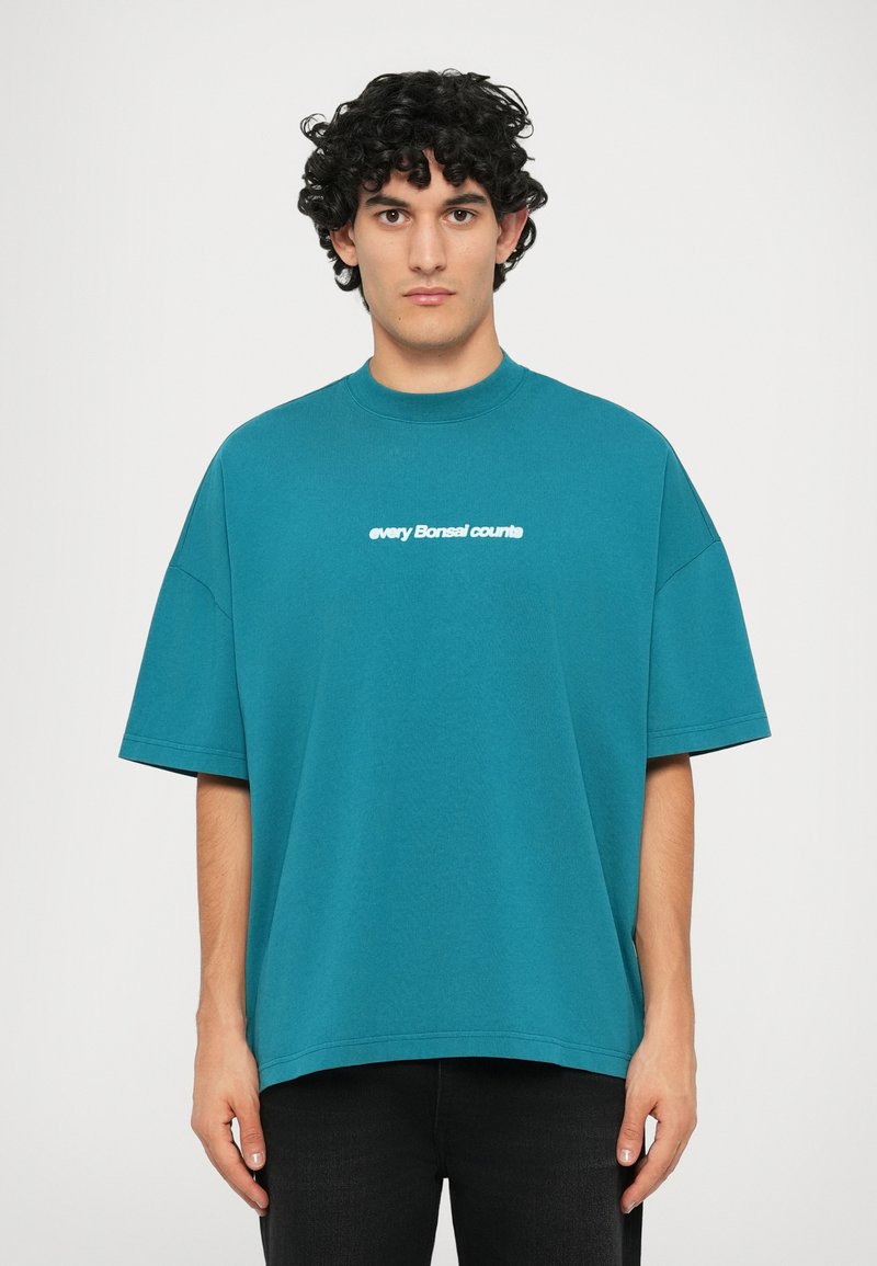 T-shirt en coton surdimensionné teal avec un col rond et des manches courtes, arborant un texte blanc "chaque Bonsai compte" sur la poitrine.
