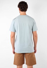 Camiseta de algodón azul claro con mangas cortas, cuello redondo y un pequeño detalle de logo en el hombro izquierdo. Combinada con pantalones cortos marrones.