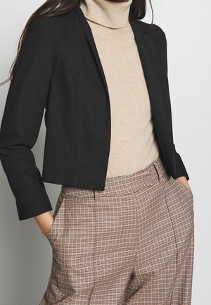 Frau trägt beigen Rollkragenpullover, schwarzen kurzen Blazer und hoch taillierte beige karierte Hose mit den Händen in den Taschen.