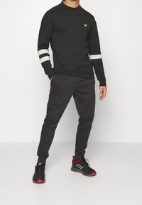 Felpa nera con dettagli a strisce bianche e un logo giallo, abbinata a pantaloni jogger neri. A completare il look ci sono scarpe da ginnastica nere e rosse.