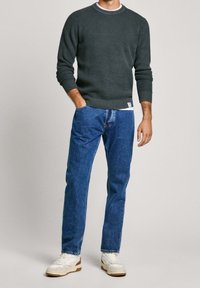 Muž v tmavě zeleném pleteném svetru, modrých džínách a bílých teniskách stojí s jednou rukou v kapse na jednoduchém pozadí.