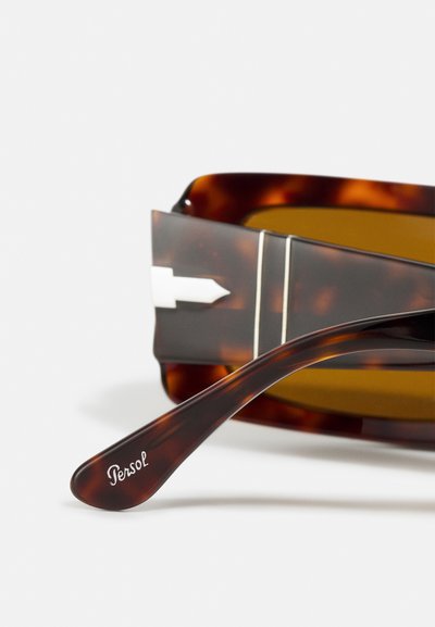 Óculos de sol tartaruga com lentes castanhas, apresentando uma dobradiça de metal e um braço curvo estampado com "Persol" em branco.