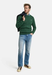 Grüner Cable-Knit-Pullover, blusehemd, hellblaue Jeans und braune Sneaker. Das Model hält eine marineblaue Jacke über der Schulter.