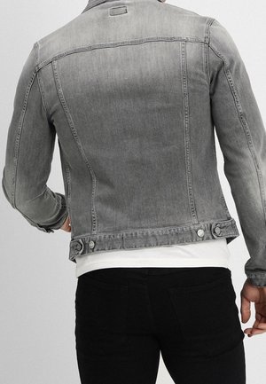 Homme portant une veste en jean grise délavée sur une chemise blanche et un pantalon noir, vu de dos sur un fond uni.