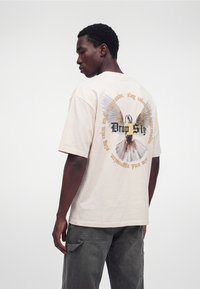 Uomo con maglietta beige chiaro con un cavallo alato e scritta circolare sul retro, abbinata a pantaloni cargo grigio scuro.