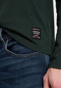 Zielona koszula z długim rękawem z naszytą czarną metką z czerwonymi akcentami z napisem "CROSS JEANS DENIM DLA ŻYCIA OD 1990", zestawiona z niebieskimi dżinsami.