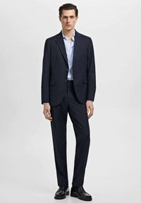 Costum bleumarin cu sacou cu un singur nasture, pantaloni asortați și cămașă albastră deschisă. Pantofii negri lucioși completează ținuta.
