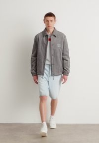 Veste en velours côtelé gris avec logo, portée sur un T-shirt blanc avec un accent rouge, associée à un short en denim clair et des baskets blanches.