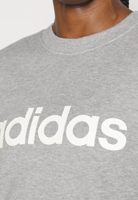 Grå sweatshirt tillverkad av mjukt tyg med en framträdande vit "adidas"-logotyp över bröstet; ribbad rund halsringning som ger extra textur.