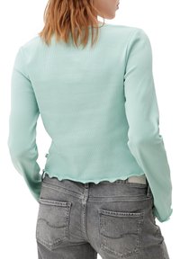 Mátově zelený žebrovaný top s dlouhým rukávem a vlnkovým lemem, spárovaný se světle šedými džíny s prošívanými zadními kapsami. Zobrazený zadní pohled.