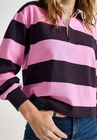 Person iført en langærmet polo-shirt med pink og sort horisontale striber, med hånden i forlommerne på blå jeans.
