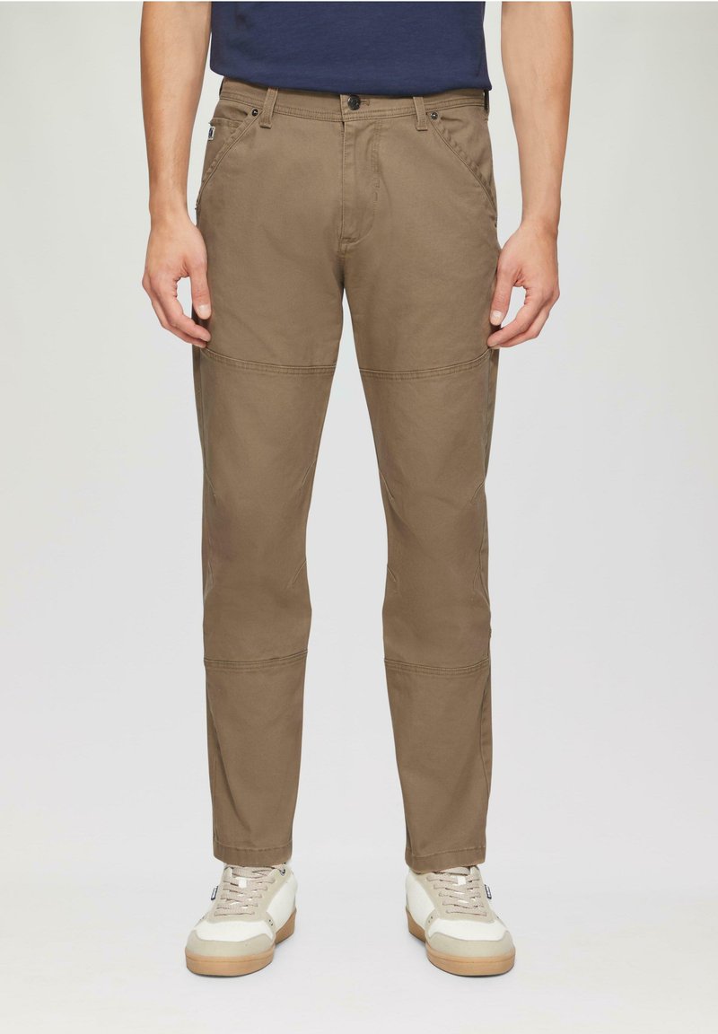 Bruine katoenen broek met een recht model, voorzien van verstevigde stiksels en een knoopsluiting. Draagt met witte sneakers.