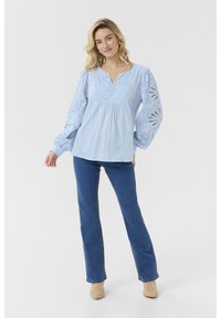 Svetlo modra bluzica z dolgimi, nabranimi rokavi, podrobnostmi iz eyeleta in V-izrezom. V kombinaciji z modrimi naravnost krojenimi kavbojkami in bež čevlji.