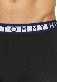 Czarne bawełniane bokserki z granatowym elastycznym paskiem, na którym widnieje napis "TOMMY HILFIGER" białymi literami w formie bloku, o gładkiej fakturze.