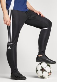 Zwarte sportieve broek met witte strepen en een Adidas-logo. De stof is glad, aansluitend, en de outfit wordt aangevuld met zwarte sportschoenen en een voetbal.