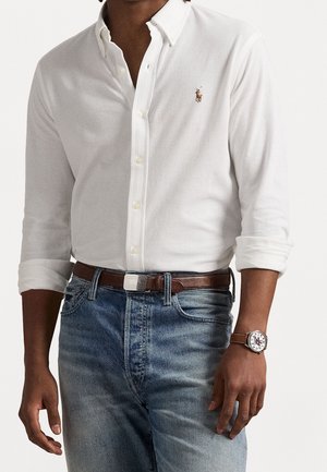 Mann mit weißem Hemd mit Knöpfen und hochgekrempelten Ärmeln, blauen Jeans, braunem Gürtel und einer Armbanduhr mit braunem Lederarmband.