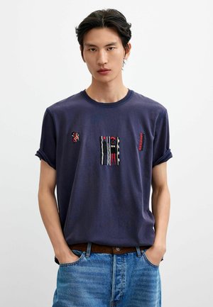 Mężczyzna o ciemnych włosach nosi granatowy t-shirt z małymi kolorowymi haftami i jasnoniebieskie dżinsy z rękami w kieszeniach.