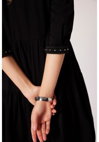 Czarna sukienka z rękawami trzyczwartowymi, z falbaniastym dołem i ozdobnymi srebrnymi ćwiekami na mankietach. Srebrna metaliczna bransoletka na nadgarstku.