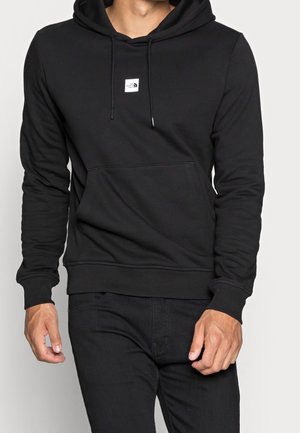 Man draagt een zwarte pullover hoodie met voorzak en een klein wit vierkant logo in het midden van de borst.